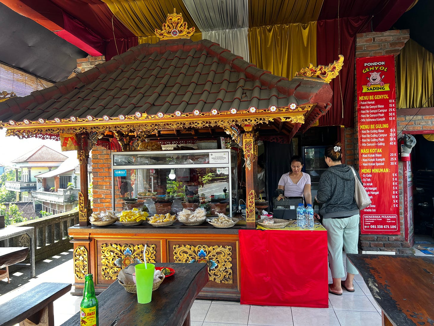 An open-air food stall with a counter carved and painted gold like a small temple, a price banner, and trays of food behind glass. 
