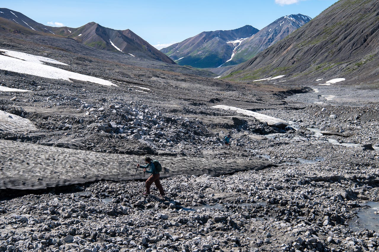 A hiker in a teal pack and brown pants crosses a broad, rubble-strewn valley floor, stepping over grey cobbles between a low band of dirty remnant snow and a braided meltwater channel. A second figure crouches on the rocks behind. Beyond, the debris-covered valley runs back to green-and-brown ridges and snow-streaked peaks under a hazy blue sky.