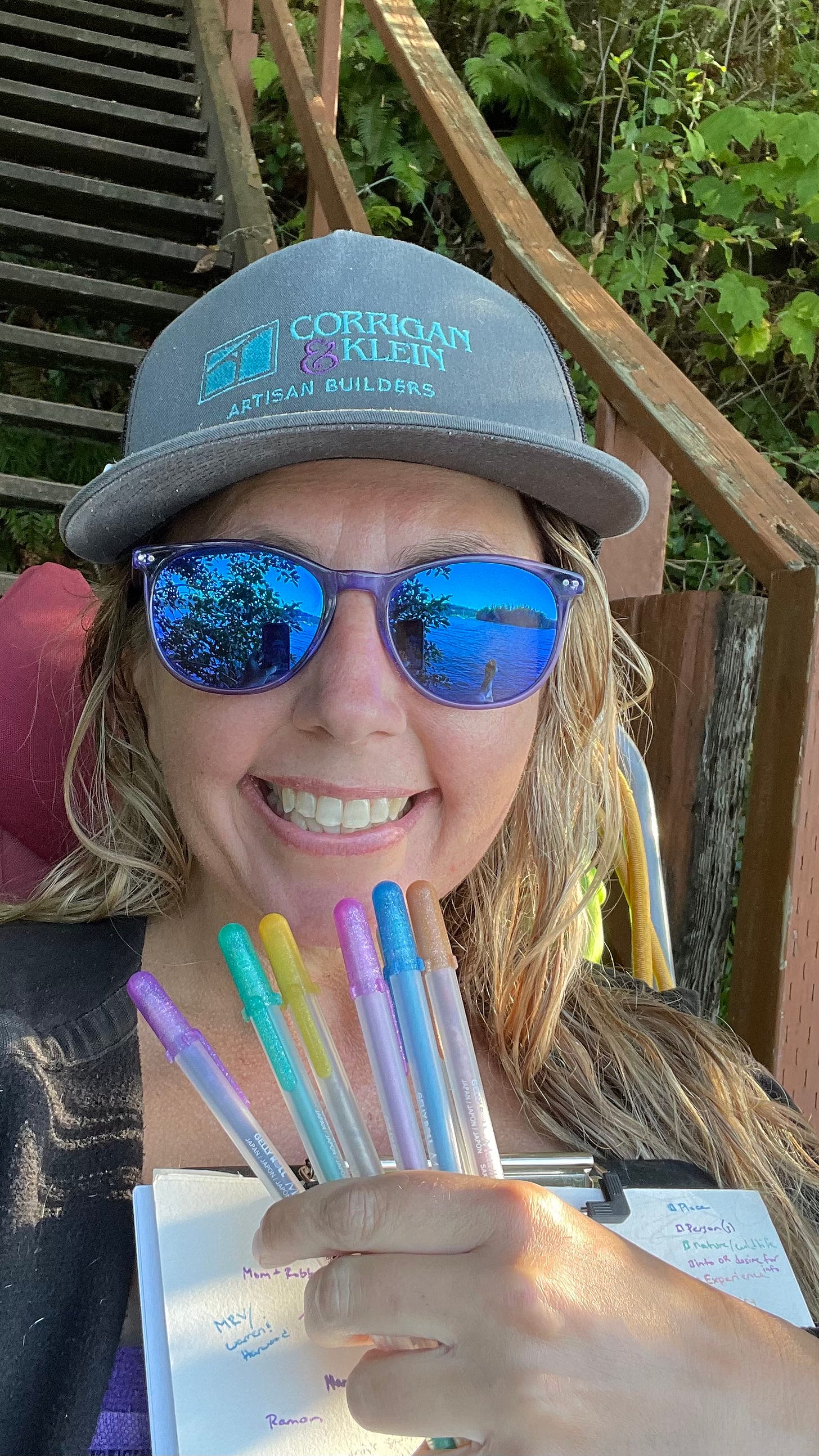 Selfie of Cassidy, a blonde white femme wearing purple sunglasses and a hat with glitter pens fanned in front of her face and a staircase in the background 