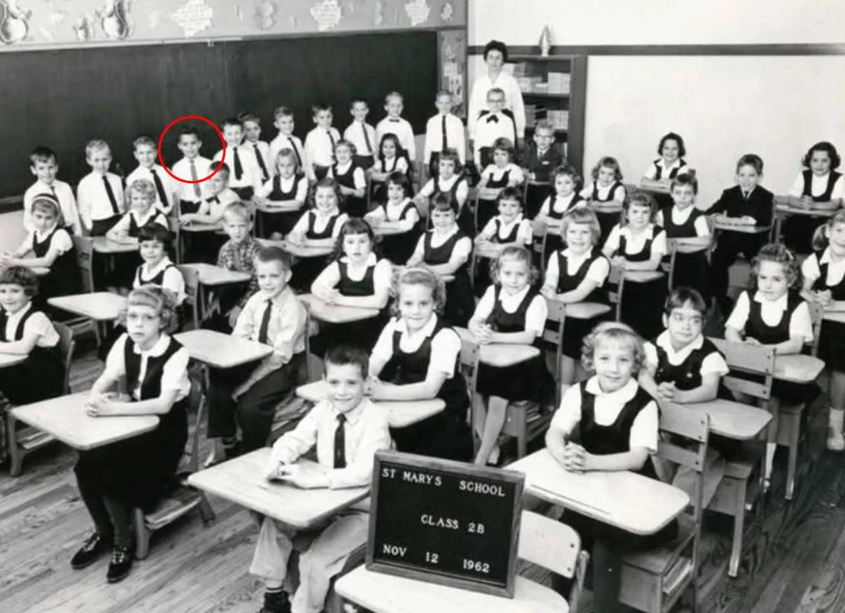 A classroom full of children pose for a picture at their desks. A sign at the front of the photo reads "St. Mary's School Class 2B Nov 12 1962
