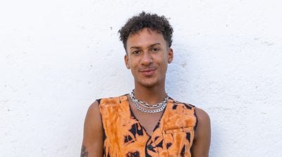 Jordon R wears a tie-dye sleeveless top in orange and black with three chunky silver chains around his neck.  He has a tattoo on his arm and some dermal face piercings