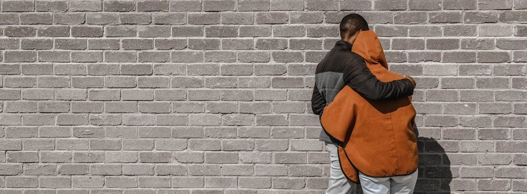 man in black and brown jacket and white pants standing beside brick wall
