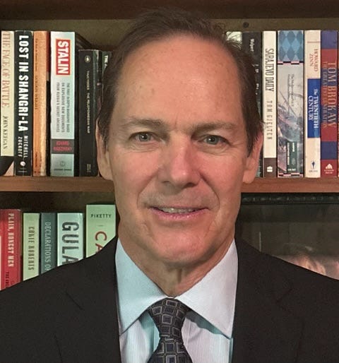 Head and shoulders of David Costello, middle-aged white man, balding, brown hair, wearing patterned tie and dark suit coat. He’s in front of a book shelf with a range of history titles, including a book on Stalin, one on the gulag, one called “Lost in Shangri La”