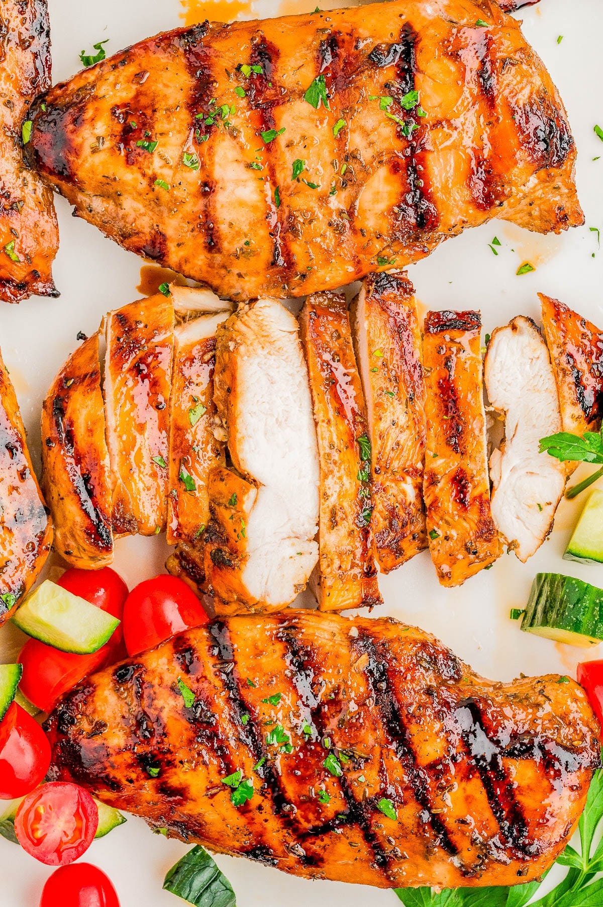 Grilled chicken breasts, some sliced, garnished with herbs and served with cherry tomatoes and cucumber pieces on a white surface.