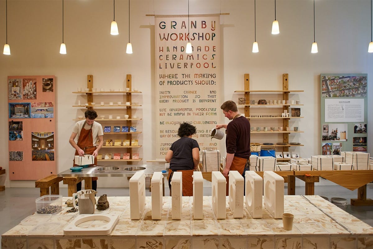 Visitors move through a working ceramics studio installed inside a gallery, where shelves, tables, clay, and handcrafted pottery demonstrate artists and community makers creating functional objects together in a shared production space.