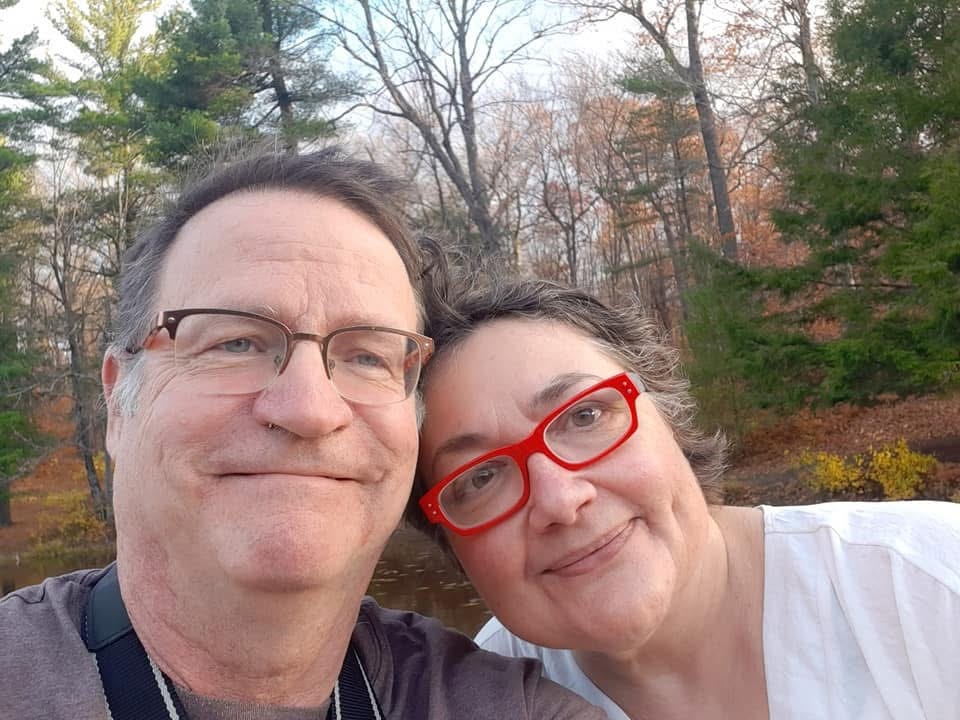 Photo of Janine's partner David and Janine, Janine's head on David's shoulder, both wearing glasses and smiling at the camera, with the spring forest in the background
