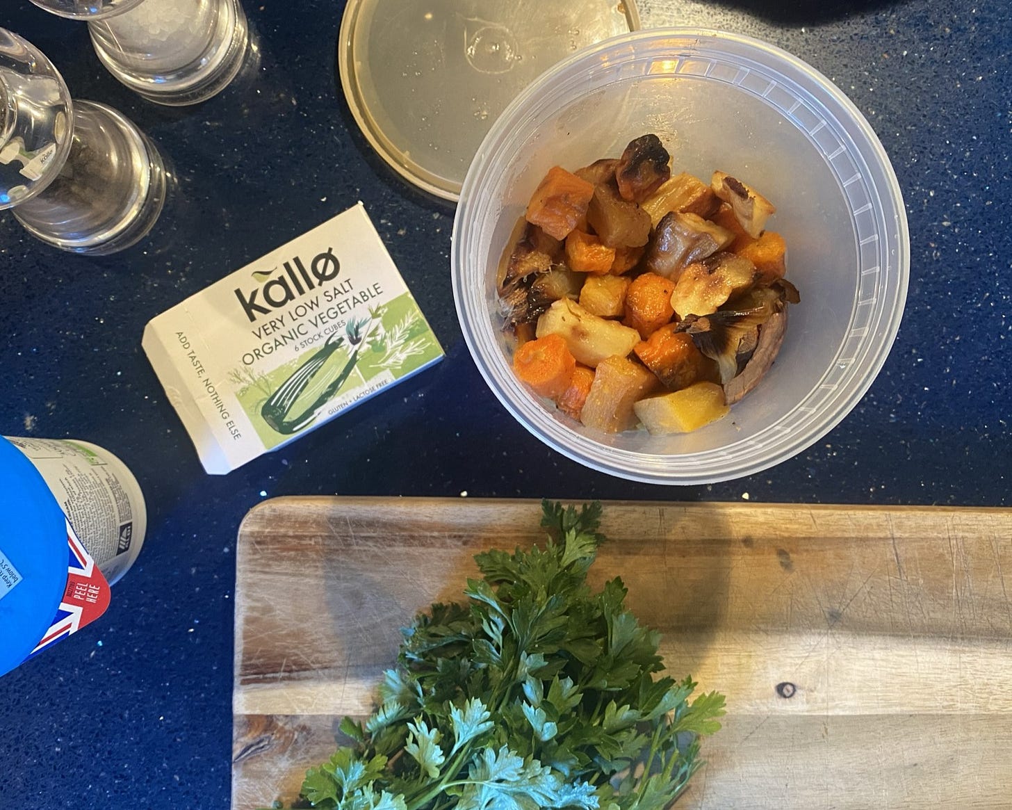Image shows a worktop and chopping board. On the board, there is a bunch of fresh herbs. On the worktop, there is a pack of Kallo stock cubes, a tub of cream, salt and pepper mills and a plastic tub with roasted vegetables