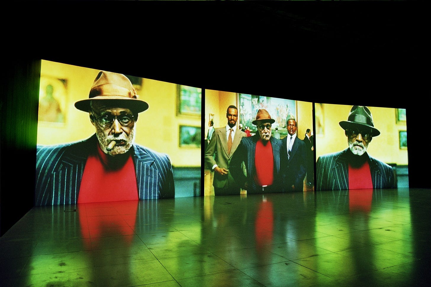 A photo of a triptych video installation titled Baltimore. All videos show the subjects in a brightly lit museum with paintings. The first video shows an elderly Black man with glasses wearing a tan fedora, red sweater, and blue/white striped blazer, with a cigar sticking out of his mouth. The middle video shows the same man flanked by wax figures of Malcolm X and Martin Luther King, Jr. The third video shows the same black man but he’s wearing a green fedora and doesn’t have a cigar in his mouth.