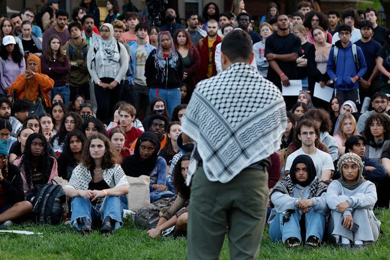 Students in a crowd (seated and standing) listen to a speaker standing in front of them. Many of them wear keffiyehs, including the speaker..