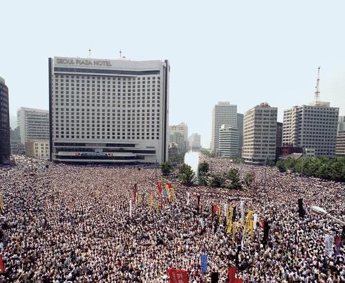 Aerial view of a vast crowd filling Seoul City Hall Plaza, Sogong-ro, and surrounding streets on July 9, 1987, during the funeral rally for student Lee Han-yeol; banners rise above people in white shirts, with the Seoul Plaza Hotel and downtown buildings in the background—a defining scene of Korea’s June Democracy Uprising.