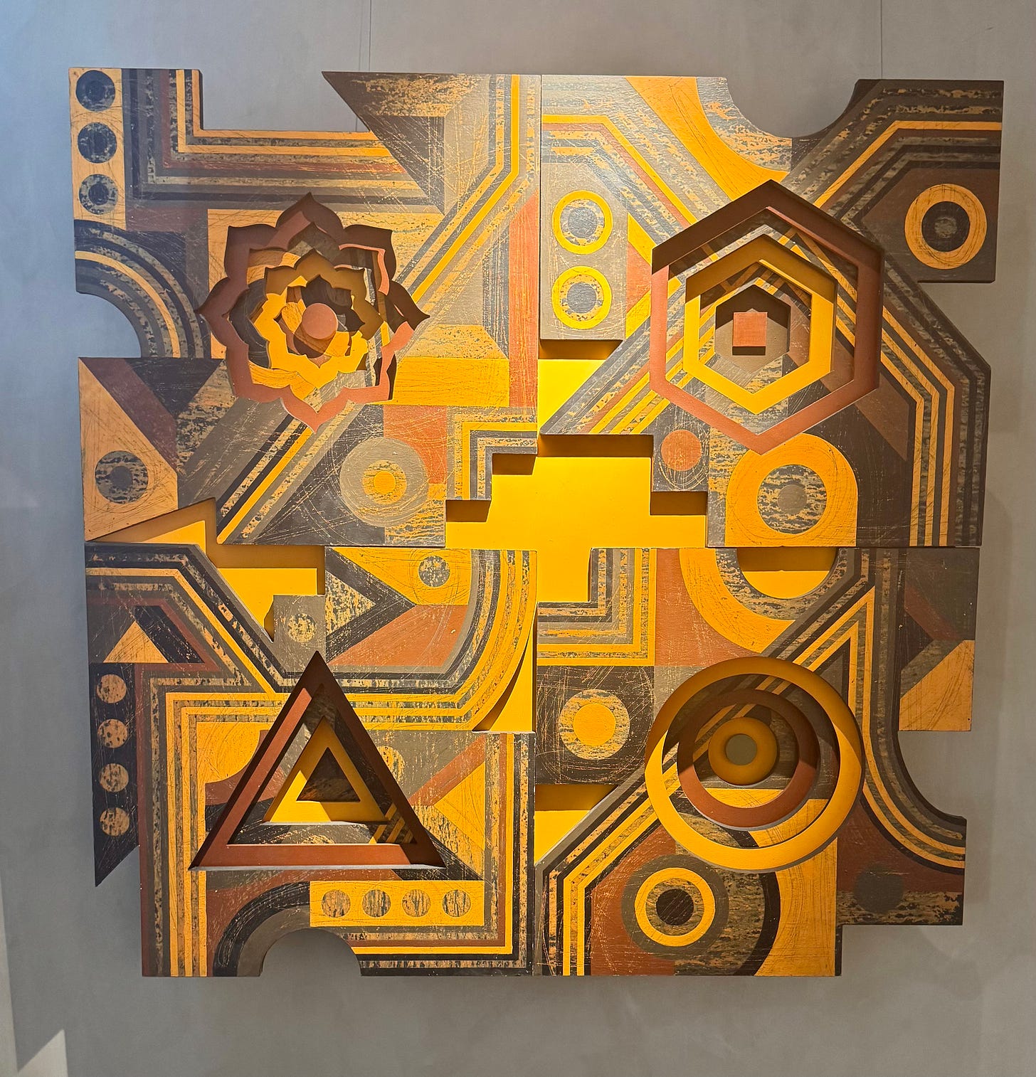 Geometric abstract artwork by Rakesh Patel in warm yellows and browns with layered symbols and architectural shapes.