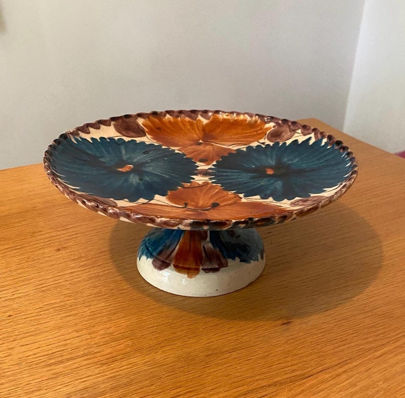 May include: A ceramic pedestal cake stand featuring a hand-painted floral pattern in deep blue and burnt orange on a cream-colored base. The circular plate has a scalloped, brown-rimmed edge, and the base is decorated with matching floral motifs.