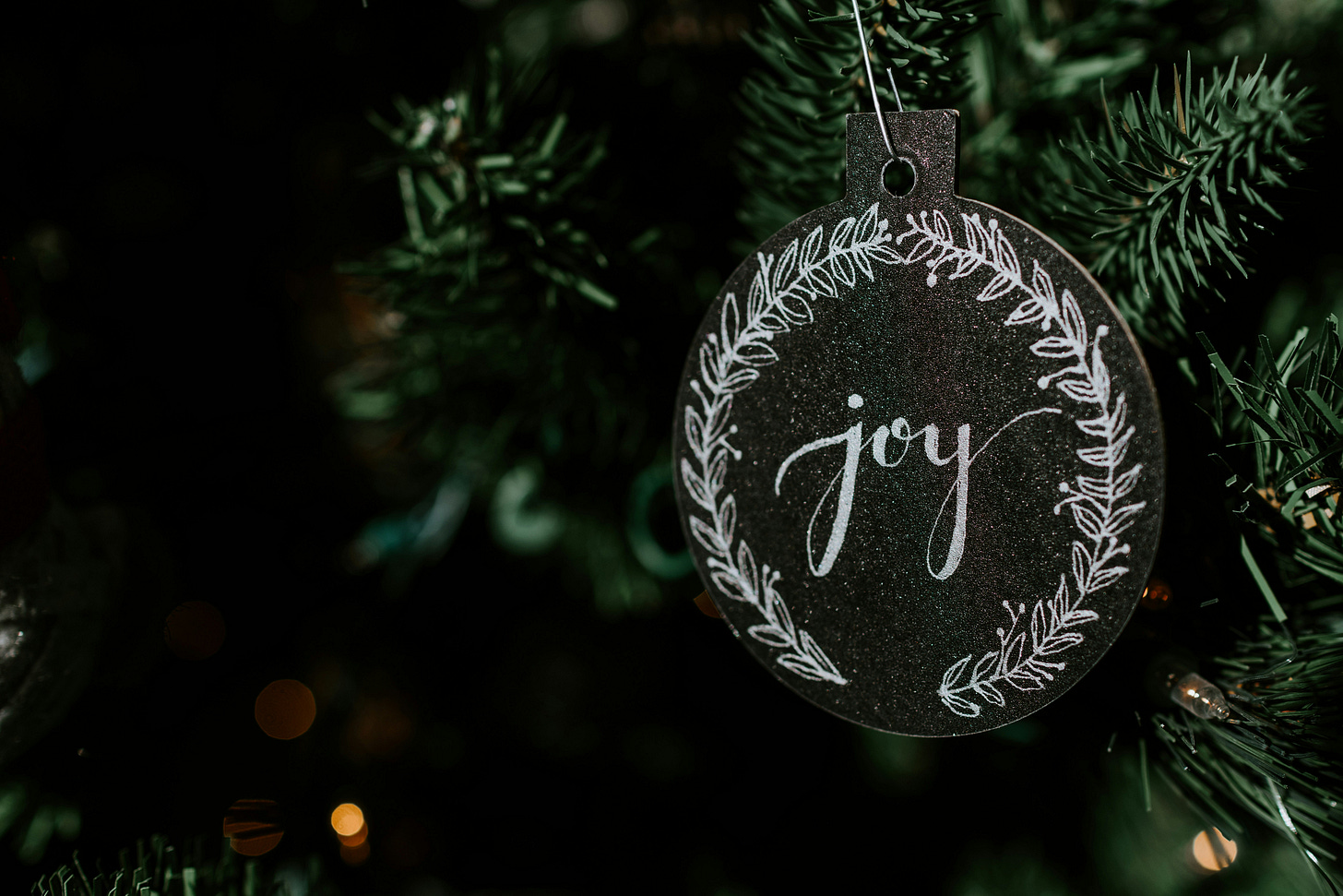Charcoal grey ornament with the word “joy” in white cursive letters on it.  There are laurels on each side of the word, “joy”. The ornament is hanging from the branch of a Christmas tree of forest green.