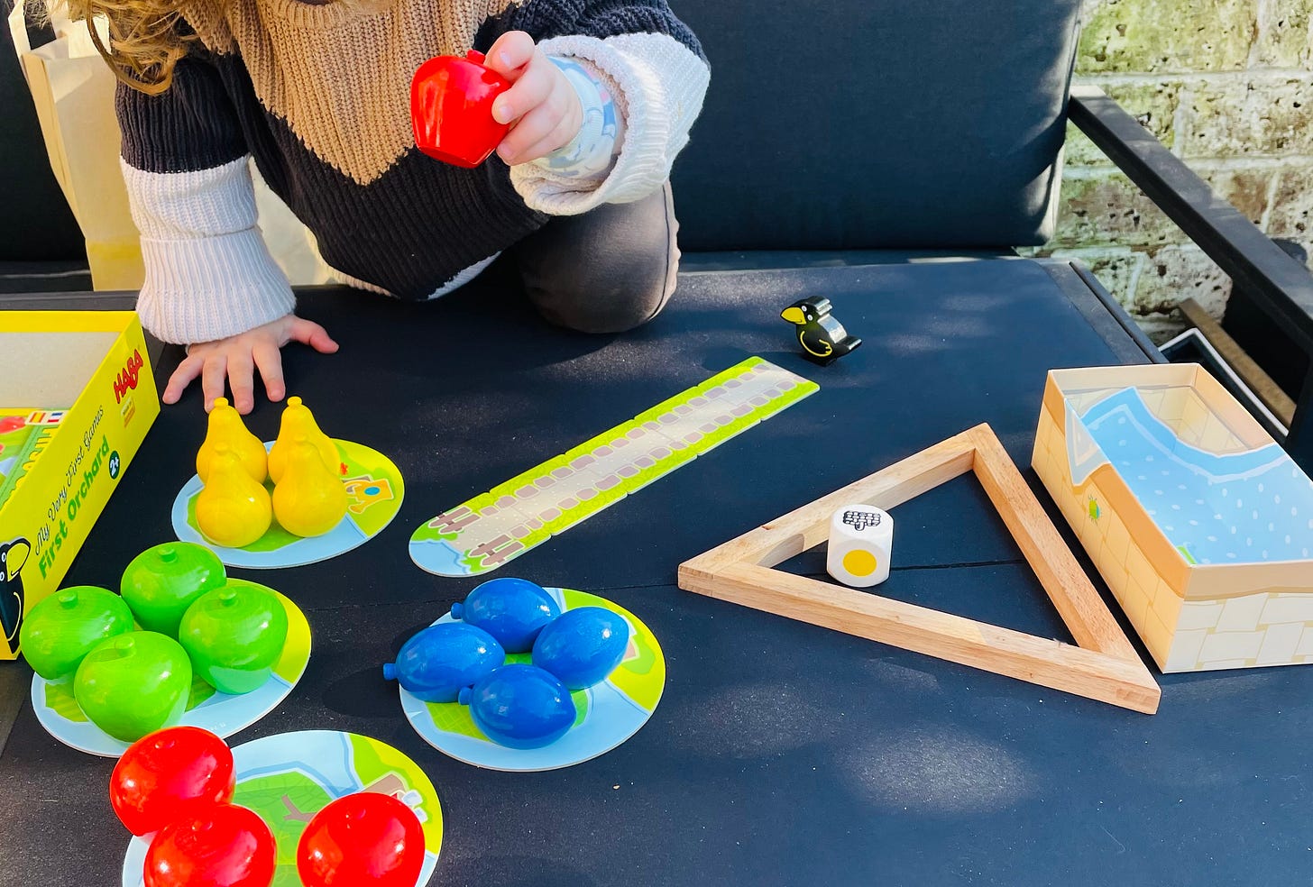 A colourful child's board game is set up on an outdoor table in the sunshine. A child is off-camera, placing a red apple piece onto the board.