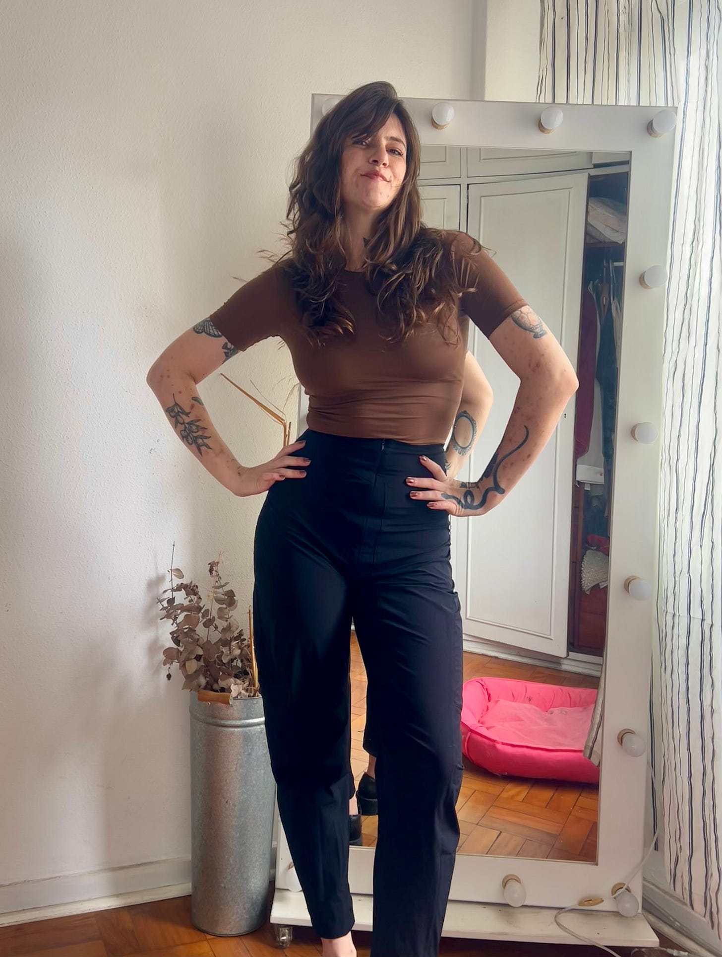 Euzinha de pé, usando uma calça preta da Insider que é mais larga nas coxas e joelhos, com uma blusa cropped de manga marronzinha bem colada no corpo