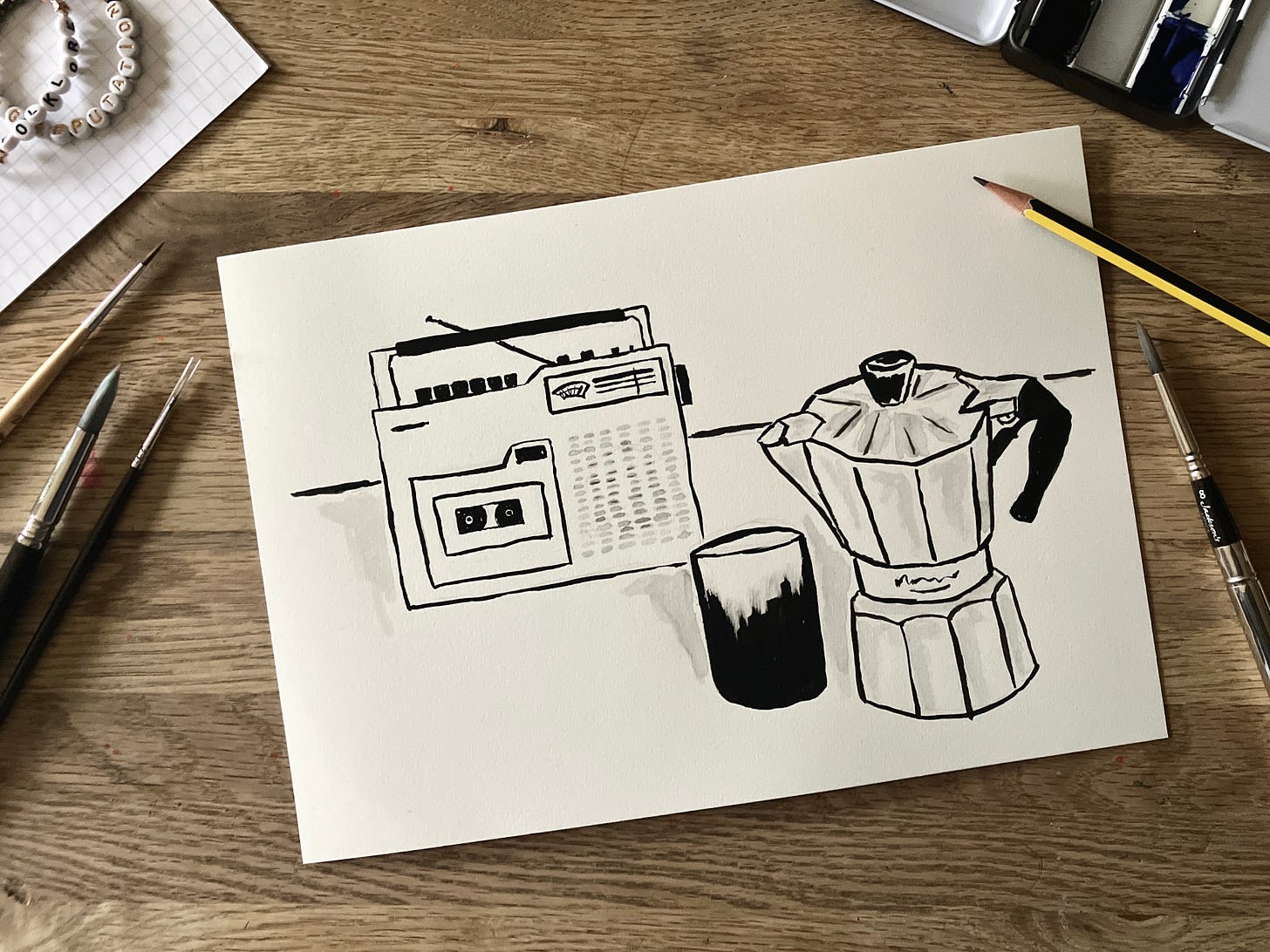 Painting of cassette player and stove top coffee maker and cup on a table sits atop a table surrounded by paint brushes and pencils