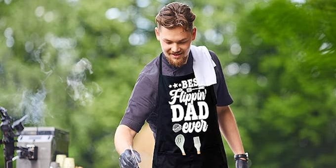 Man smiling while grilling outdoors, wearing a black apron that reads "Best Flippin' Dad Ever." Find great Father's Day gifts for the meal prep Dad in your life, like this APASIN Cooking Apron for Dad, Grilling apron for men with 2 large pockets, Adjustable neck strap, Gift for Dad, Father