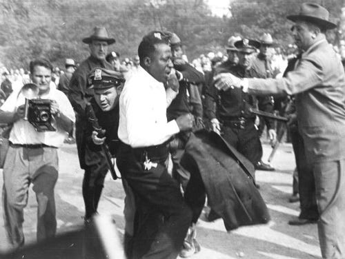 Black man looking stunned, surrounded by white news photographers and armed police