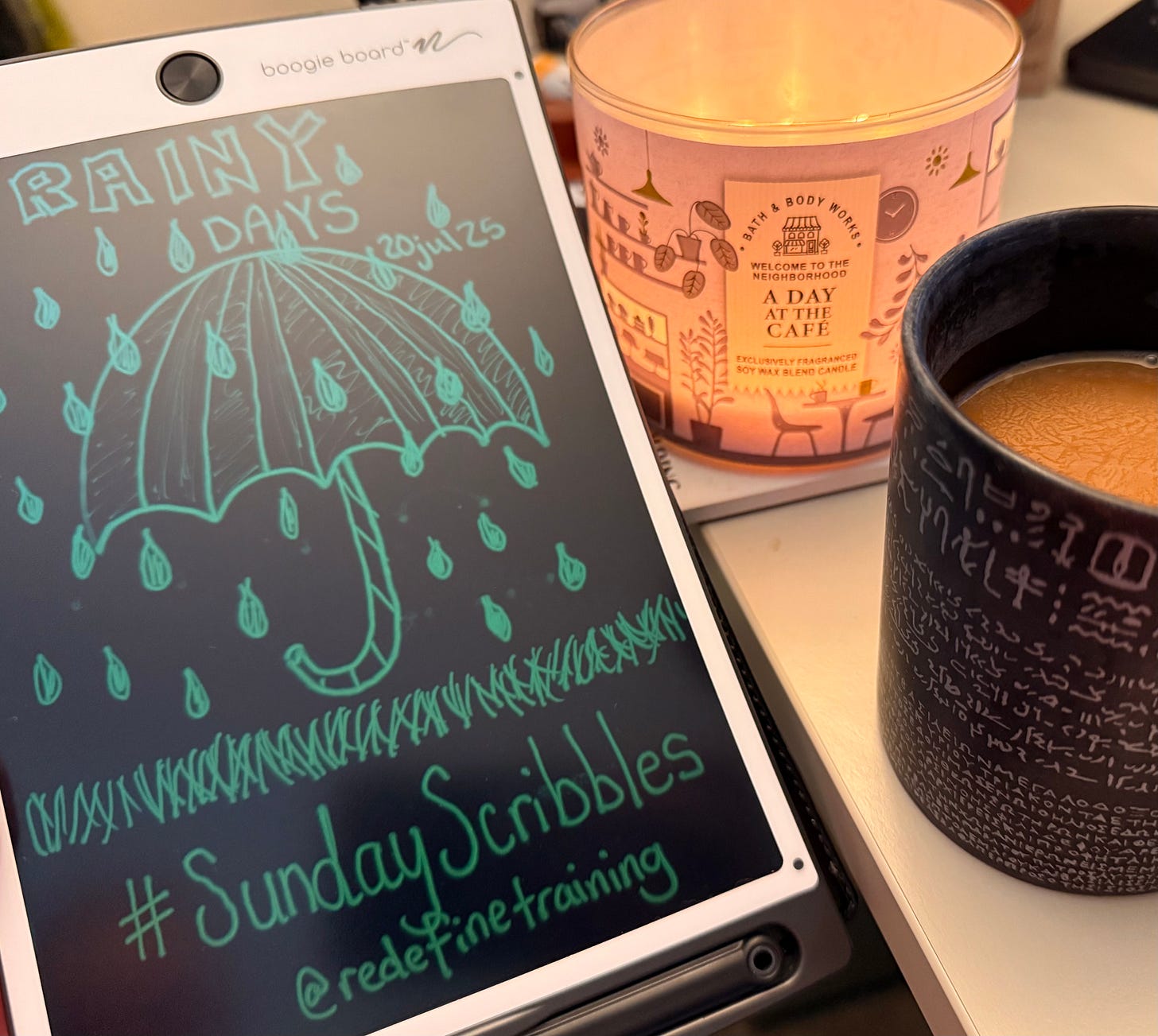 A cozy scene showing a digital writing tablet with a hand-drawn scribble of an umbrella in the rain and the hashtag #SundayScribbles. Beside it is a lit candle and a warm mug of coffee.