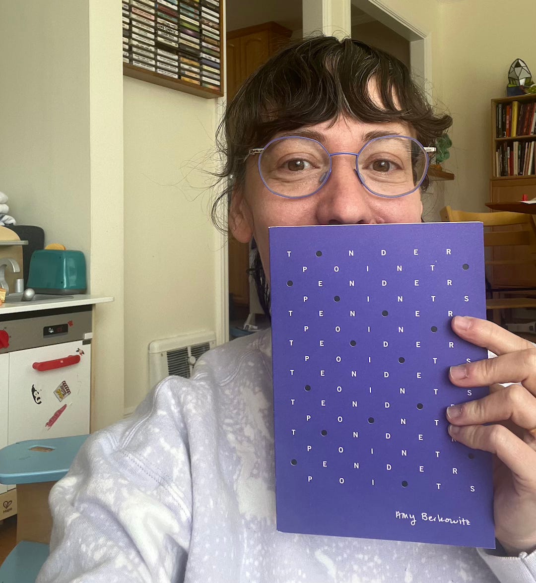 Me wearing a lavender bleach-dyed sweater and my blueish purple glasses and holding my purple book. Lots of purple. My kid’s play kitchen and our cassette tape collection are behind me. 