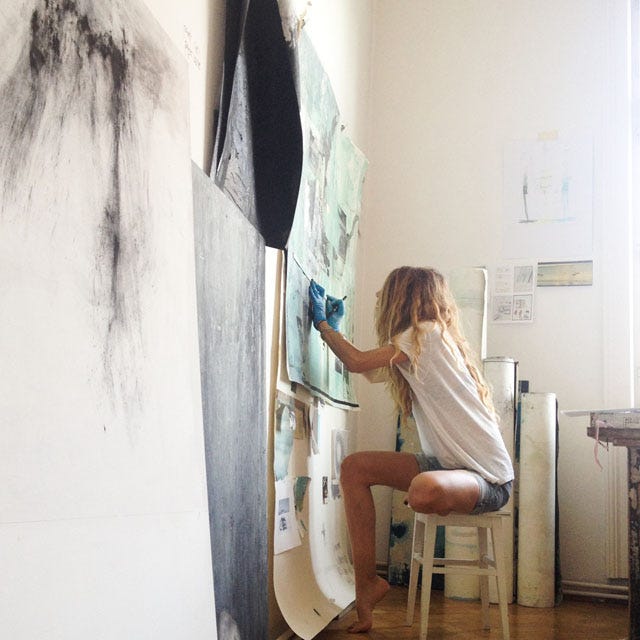 Side portrait of a woman with blonde curly hair with gloves on, sitting on a stool, drawing with charcoal in an artist studio Side portrait of a woman with blonde curly hair with gloves on, sitting on a stool, drawing with charcoal in an artist studio