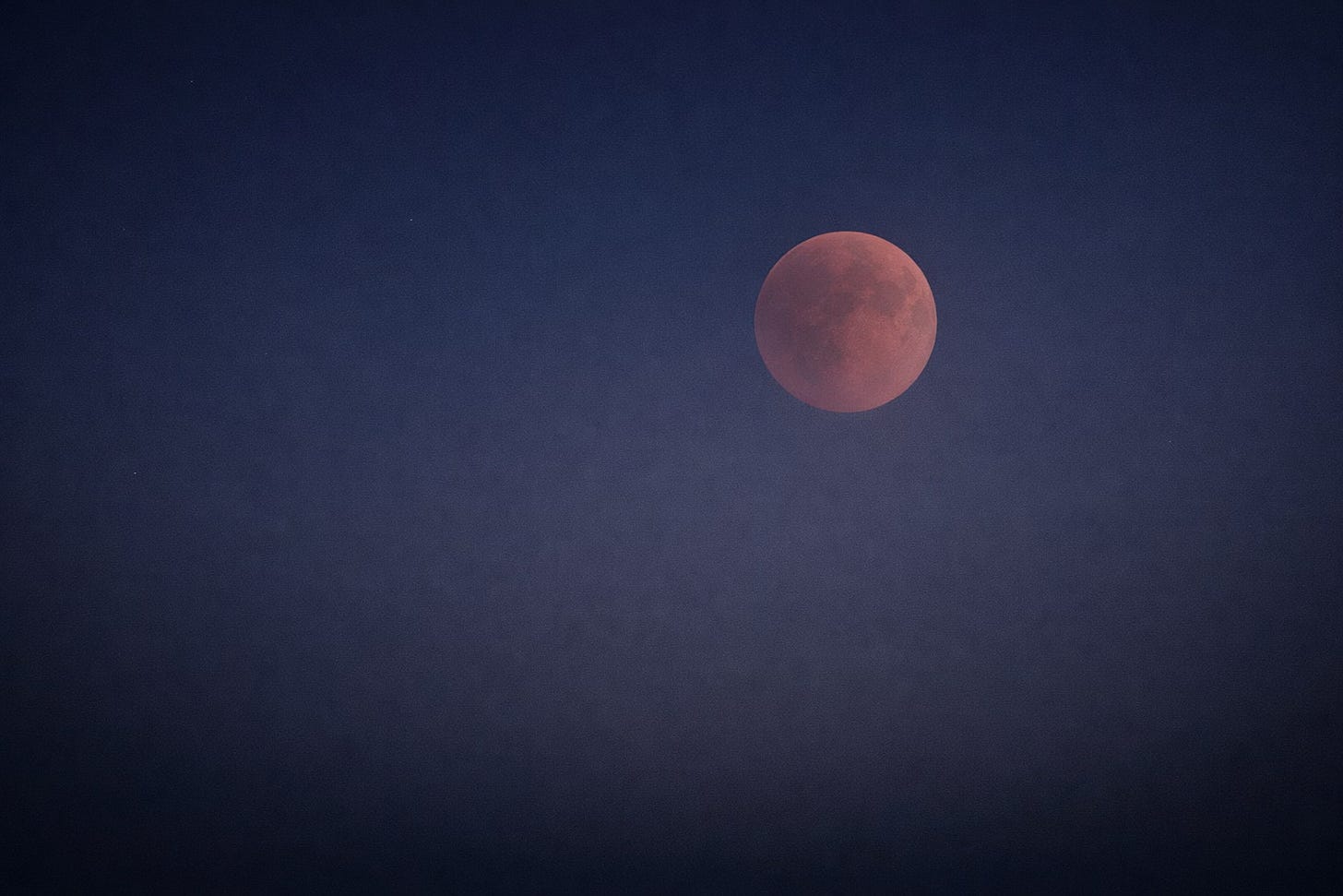 A total lunar eclipse glowing muted copper-red in a deep indigo night sky, softened by atmospheric haze and surrounded by wide, quiet negative space.