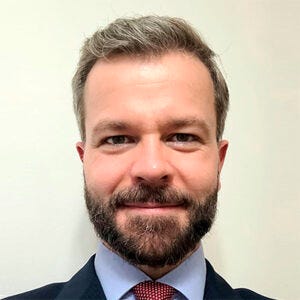 Retrato de um homem adulto, de frente para a câmera, com cabelos curtos grisalhos e barba aparada. Veste terno escuro, camisa clara e gravata vermelha. Apresenta expressão neutra com leve sorriso. O fundo é liso e claro Retrato de um homem adulto, de frente para a câmera, com cabelos curtos grisalhos e barba aparada. Veste terno escuro, camisa clara e gravata vermelha. Apresenta expressão neutra com leve sorriso. O fundo é liso e claro