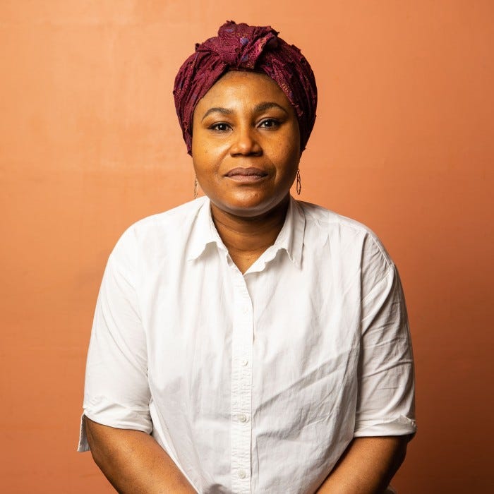 A portrait of a woman dressed in a crisp white shirt and intricate maroon headwrap, seated against an orange backdrop with a composed demeanor