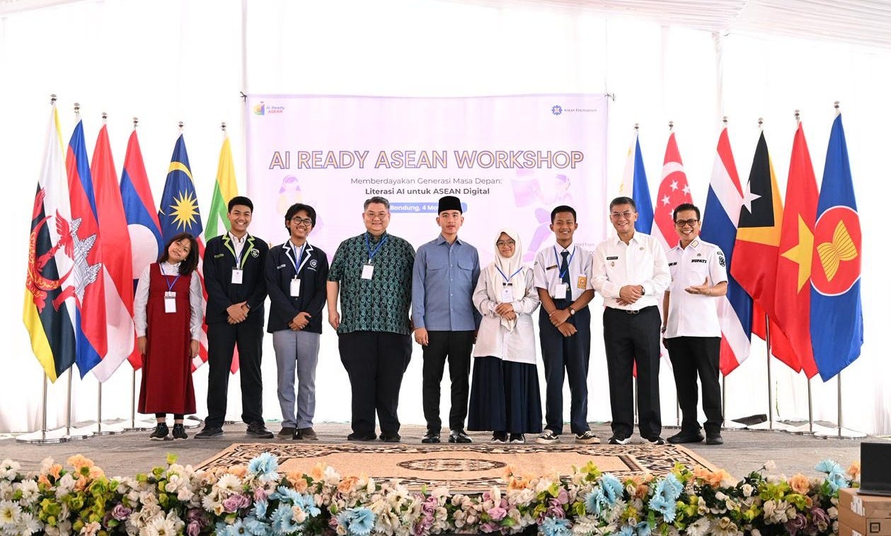 A group photograph shows ten people standing on a stage during an "AI READY ASEAN WORKSHOP." They are arranged in a line with men and young students. A banner hangs above them, reading, "AI READY ASEAN WORKSHOP" and "Memberdayakan Generasi Masa Depan: Literasi AI untuk ASEAN Digital." There are also details regarding the workshop's date and location. National flags from various ASEAN countries are displayed prominently on the stage. Below the stage, there's a dense arrangement of colorful flowers and laptops on the floor.