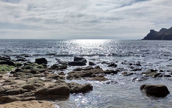 A photograph of the transition from a rocky beach to the sea, with sunlight reflecting on the water and the end of a headland in the distance on the right of the image.