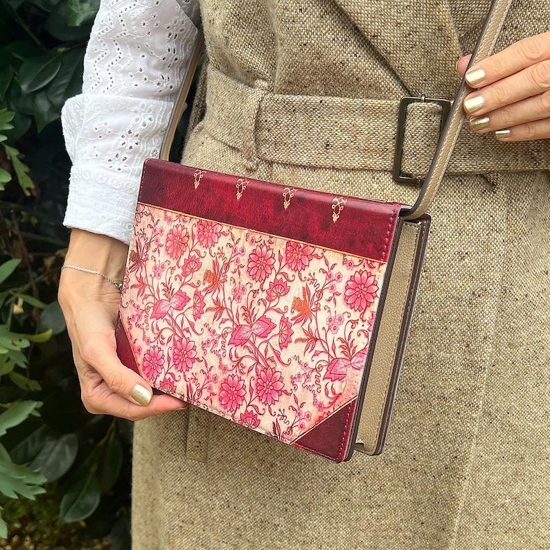 May include: A red and beige floral book clutch purse. The clutch has a beige background with a repeating pink floral pattern. The clutch has a red border and gold accents. The clutch is shown closed. The clutch has a long beige strap.