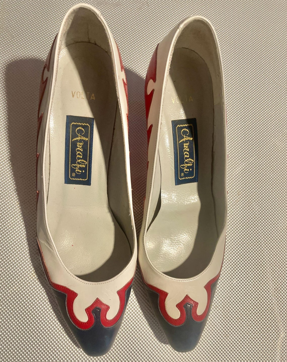 May include: A pair of white leather heeled shoes with red and blue accents. The shoes have a pointed toe and a small heel. The inside of the shoes are white with the words 'Volta' and 'Cavaldi' printed on the insole.