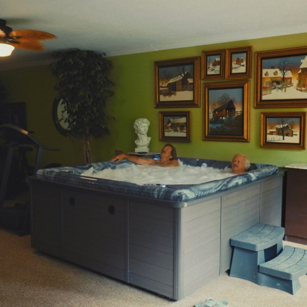 A still from the show Neighbors, showing two elderly gay men relaxing in their hot tub which is in the middle of a living room, I think? The walls are a horrible shade of green and covered in framed paintings, and I just don't understand the layout of their house