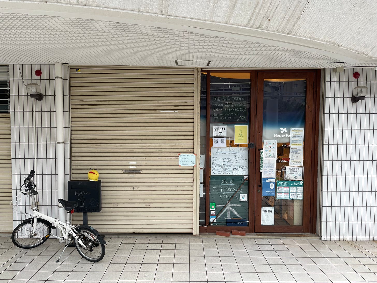 早朝の本屋lighthouse店外。シャッターが半分閉まっていて、ポストの上に温泉マークのパペットが乗っている。