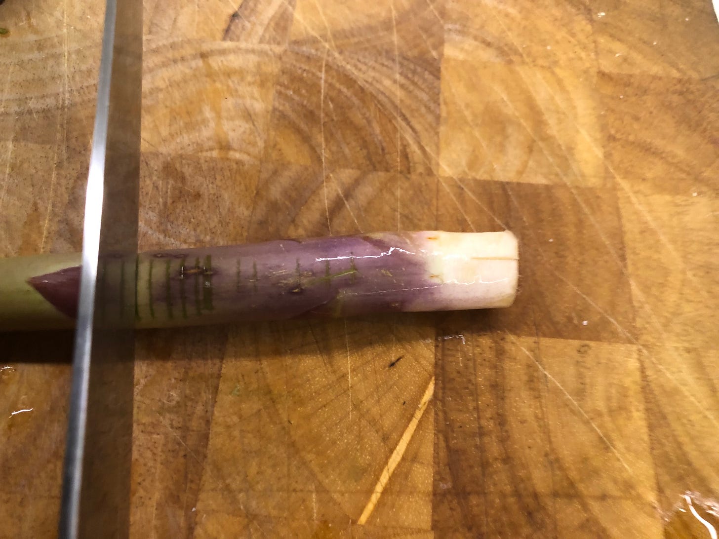 A spear of asparagus on a cutting board. It's stem is marked with several shallow cuts. A knife has cut more deeply into it farther away from the woody end. 