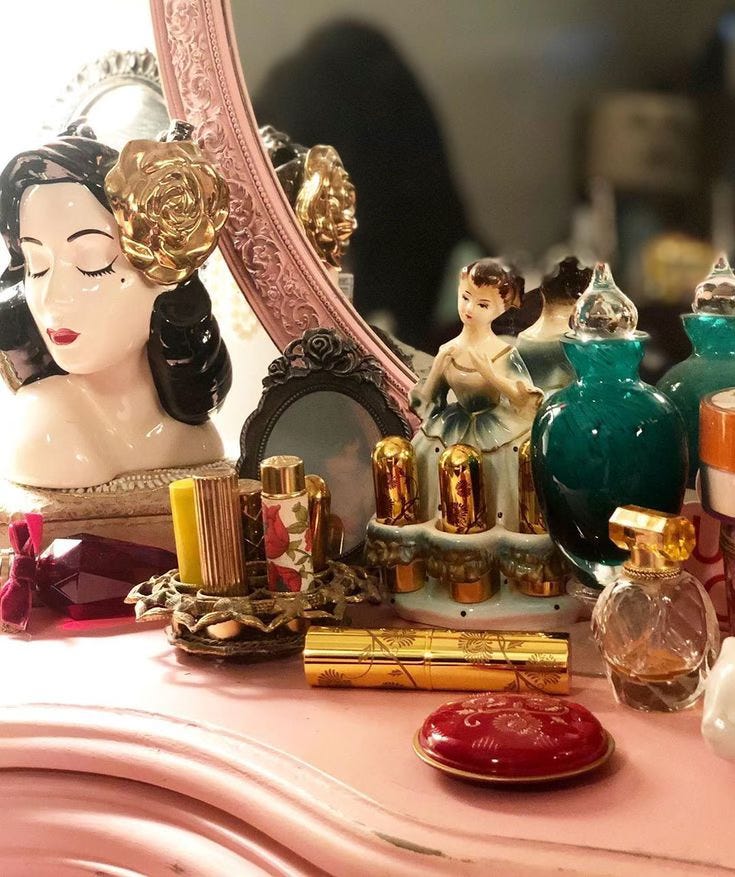 A vintage vanity setup featuring a porcelain bust with a golden rose hair accessory, ornate perfume bottles, gold and floral lipstick tubes, and a ballerina figurine. A vintage vanity setup featuring a porcelain bust with a golden rose hair accessory, ornate perfume bottles, gold and floral lipstick tubes, and a ballerina figurine.