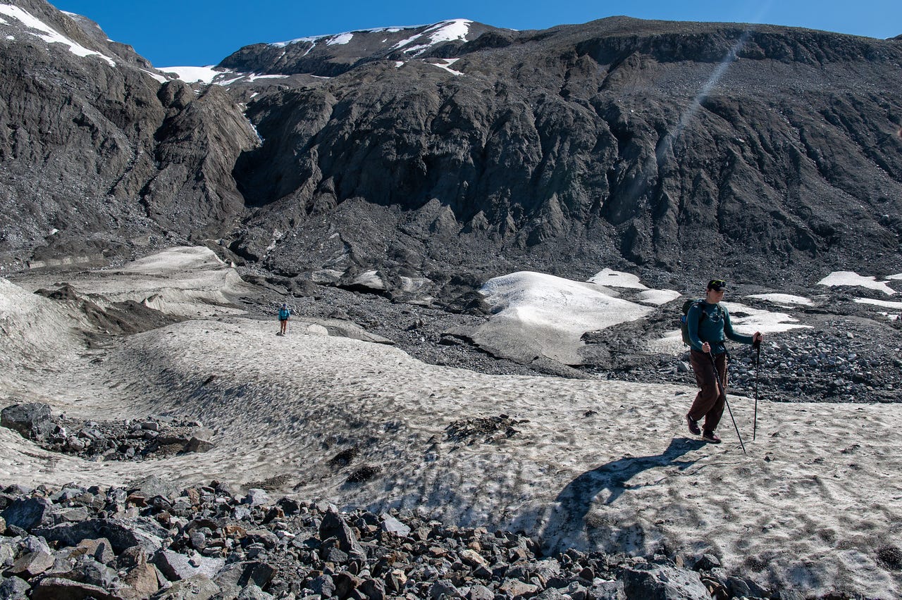 A hiker in a teal top and brown pants strides across a broad expanse of dirty, sun-cupped snow, trekking pole in hand and casting a long shadow. A second, smaller figure crosses the snow further back at left. Behind them a tall, near-black wall of ice-cored moraine rises, furrowed and streaked and patched with old snow along its top, under a deep blue sky.