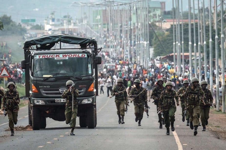 Men wearing military fatigues and helmets and holding teargas guns run ahead of a crowd of civilians, with a military truck keeping pace with the officers. Men wearing military fatigues and helmets and holding teargas guns run ahead of a crowd of civilians, with a military truck keeping pace with the officers.
