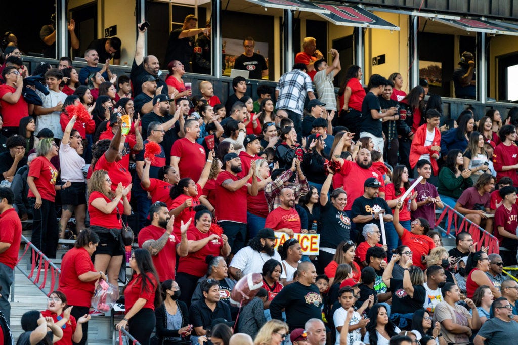 Color photograph of standing, cheering fans in the stands at a high school football game. The photo shows about twelve row of fans, mostly standing in full bleachers, cheering with arms raised, many wearing red.