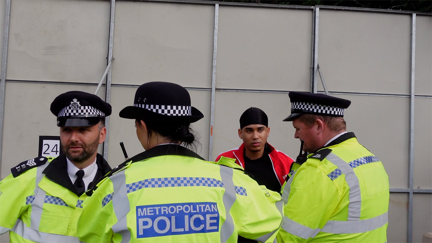 Three Metropolitan Police officers speaking with Joshua Arthur beside metal hoarding in London, September 2021. Three Metropolitan Police officers speaking with Joshua Arthur beside metal hoarding in London, September 2021.