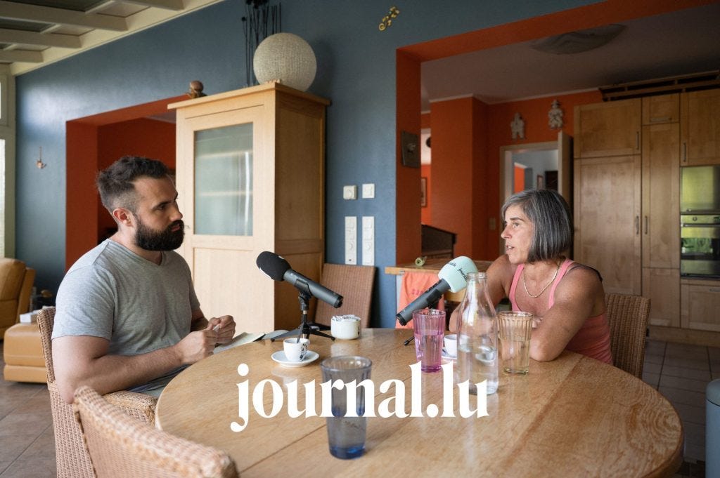 Two people sit at a round wooden table in a home kitchen and dining area, recording an interview with tabletop microphones, glasses, coffee cups, and a water bottle between them.