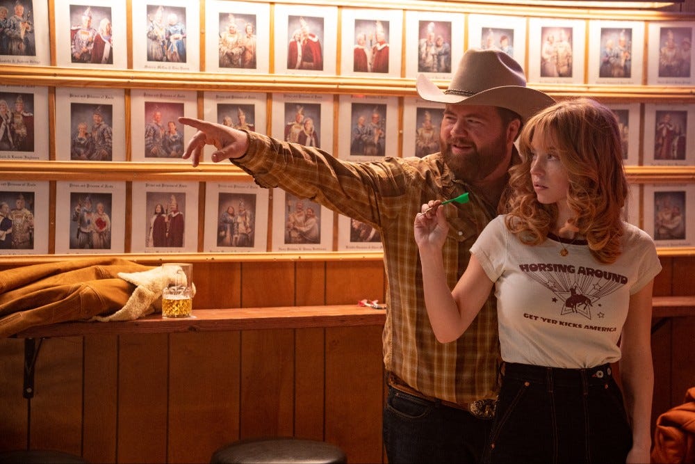 Paul Walter Hauser directing Sydney Sweeney how to throw a dart in Americana
