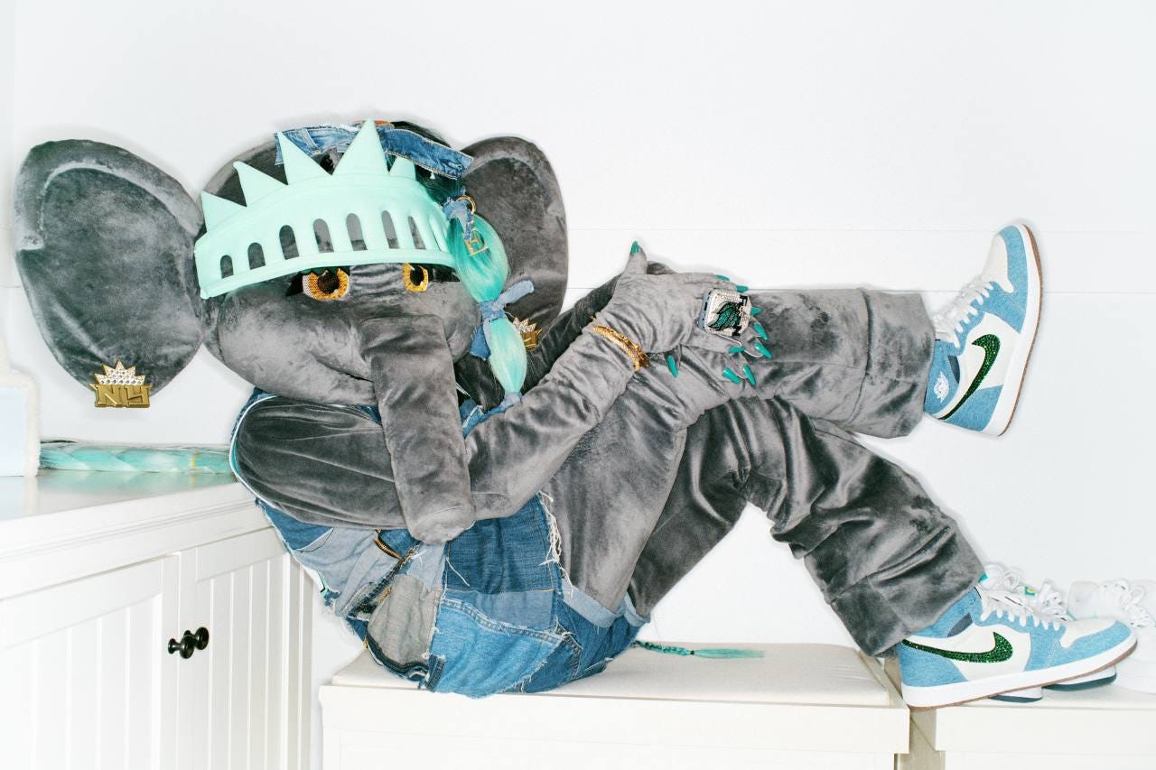 Ellie the Elephant, a female fuzzy gray elephant mascot, posing in a Statue of Liberty crown, denim clothes and blue and green Nike shoes. 