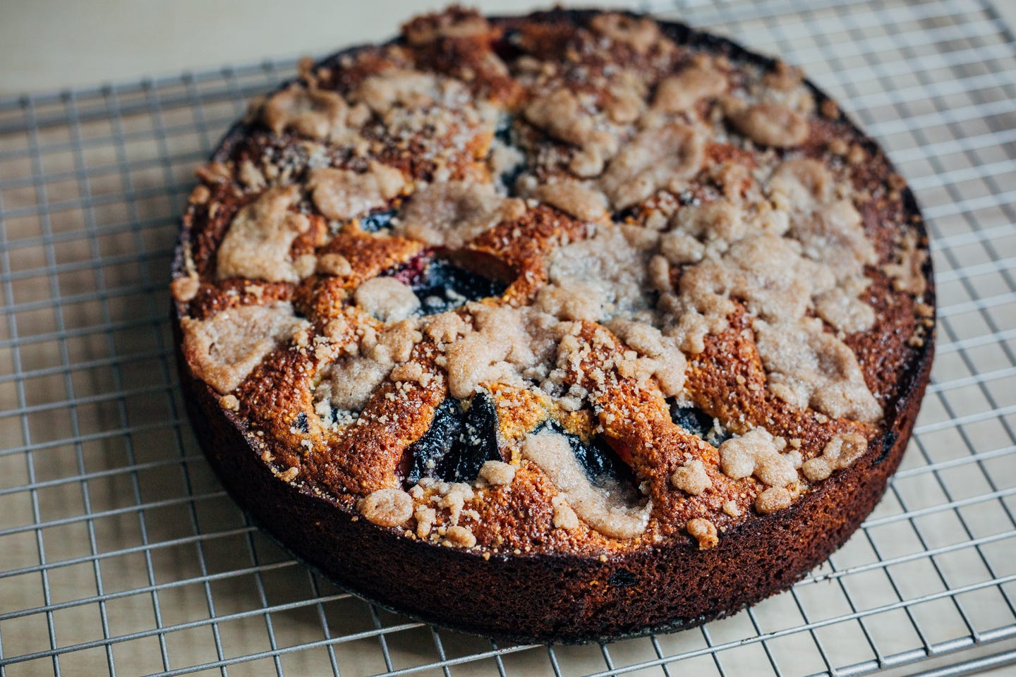 A cake cool on a wire rack. It has a golden topping and hlaved plums peaking through. 