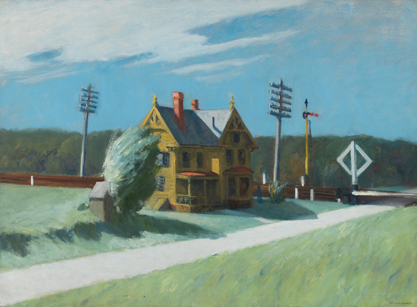 A railroad track crosses a country road in front of a house and a line of trees.
