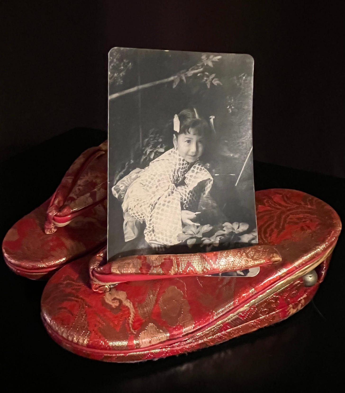 A photo of red getas, fancy traditional Japanese sandal-like shoes, and of a photograph now tucked into one of the getas of the author about 3 ½ years old, wearing a kimono, crouched on a large rock and smiling. 