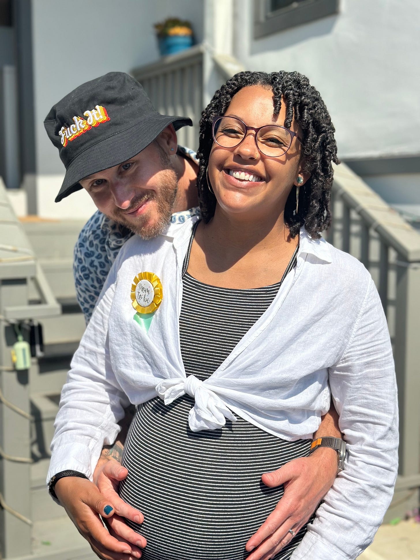 The author, wearing a bucket hat that says "Fuck it," with his arms wrapped around his very pregnant wife.