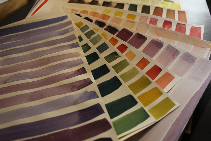 Watercolor practice sheets with rows of color swatches in purple, green, orange, and red tones stacked on a desk. Close-up of watercolor practice sheets showing strips of purple, green, yellow, and red color swatches arranged diagonally.