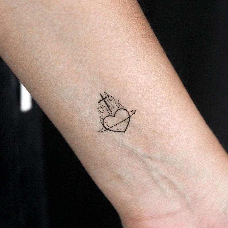 May include: A temporary tattoo of a sacred heart pierced by an arrow and engulfed in flames, with a cross on top. The design is simple, black ink on a white background. The tattoo is small and delicate, suitable for wrist placement.