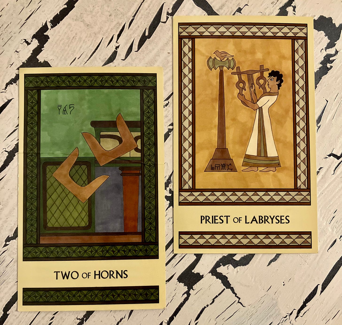 Two Minoan Tarot cards side by side on a crackled cream painted wood surface. Both cards have intricate borders in the same color family as the rest of the card. The Two of Horns is painted in shades of deep green and brown. It shows two pairs of Minoan sacred horns in midair as they tumble off a shrine. The Priest of Labryses is painted in shades of gold, cream, and tan. It shows a Minoan man with short hair, wearing a long robe and playing a large lyre. He stands facing left. On his left is a large labrys that's taller than he is, topped by a perching bird. 